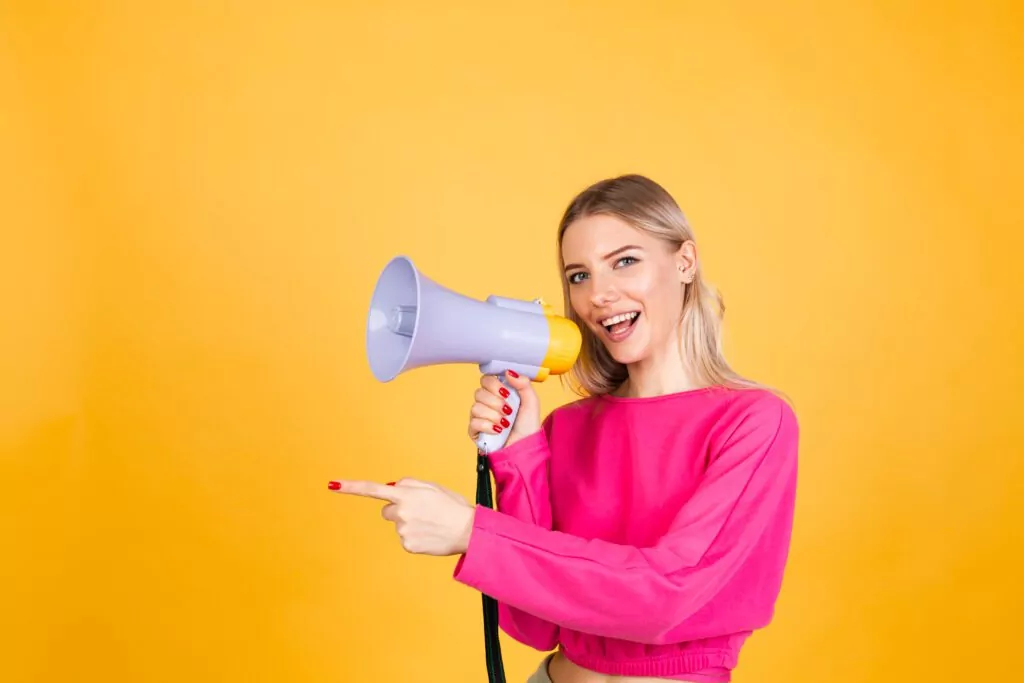 Pretty european woman in pink blouse on yellow background. Shouting in megaphone and pointing as a call to action.